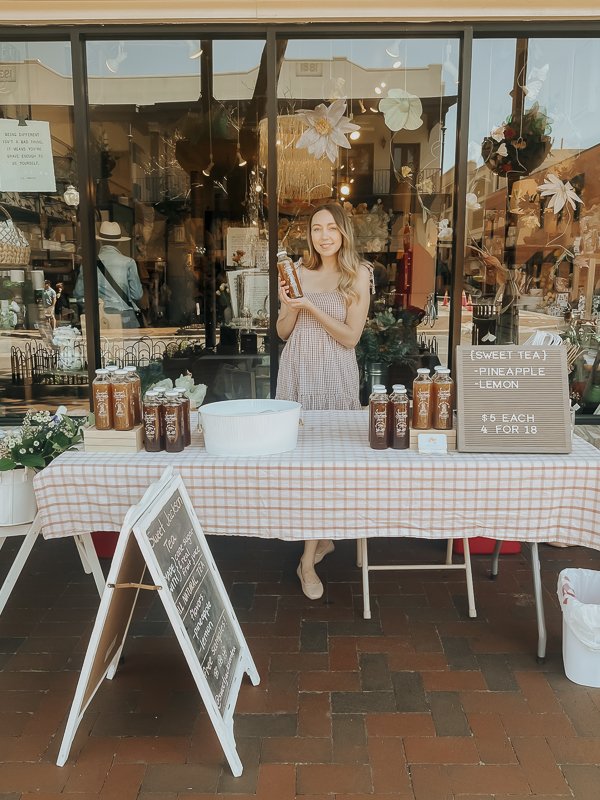 Smiling woman standing behind table holding up glass bottled sweet tea | Sweet Jackson Tea in Pensacola