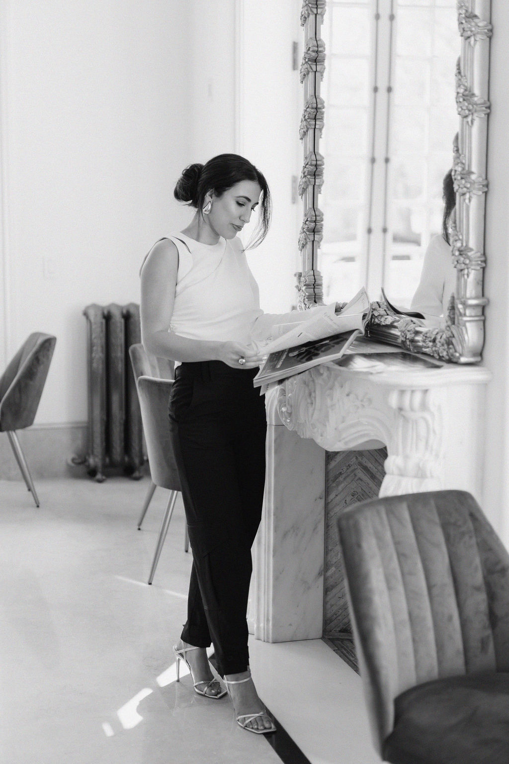 Woman flips through a magazine while leaning against an ornate mantelpiece under a large mirror | By Sofia Brown