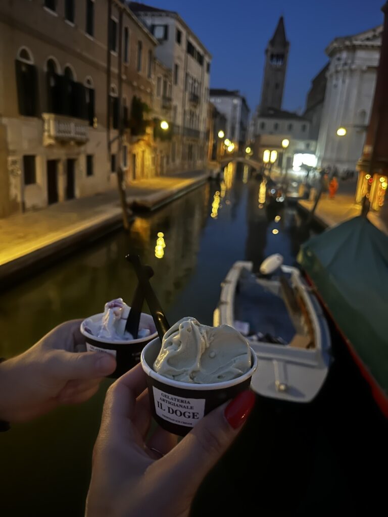 Two hands hold small cups of gelato in front of a canal in Venice at night time