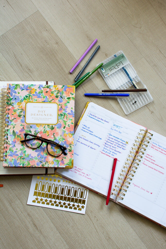 Day Designer planner sits open next to a second floral print Day Designer planner next to stickers, pens, and reading glasses