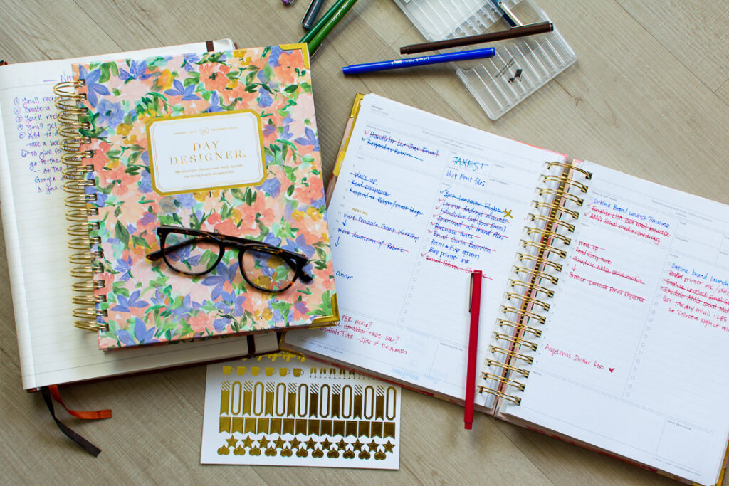 Day Designer planner sits open next to a second floral print Day Designer planner next to stickers, pens, and reading glasses