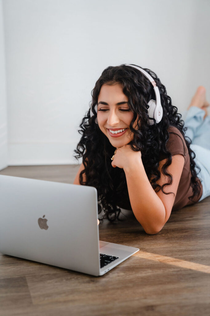 Woman with dark curly hair wearing a brown shirt, jeans, and Beats headphones lays on the floor and smiles while typing on her laptop | email marketing best practices for small business owners blog by Sofia Brown