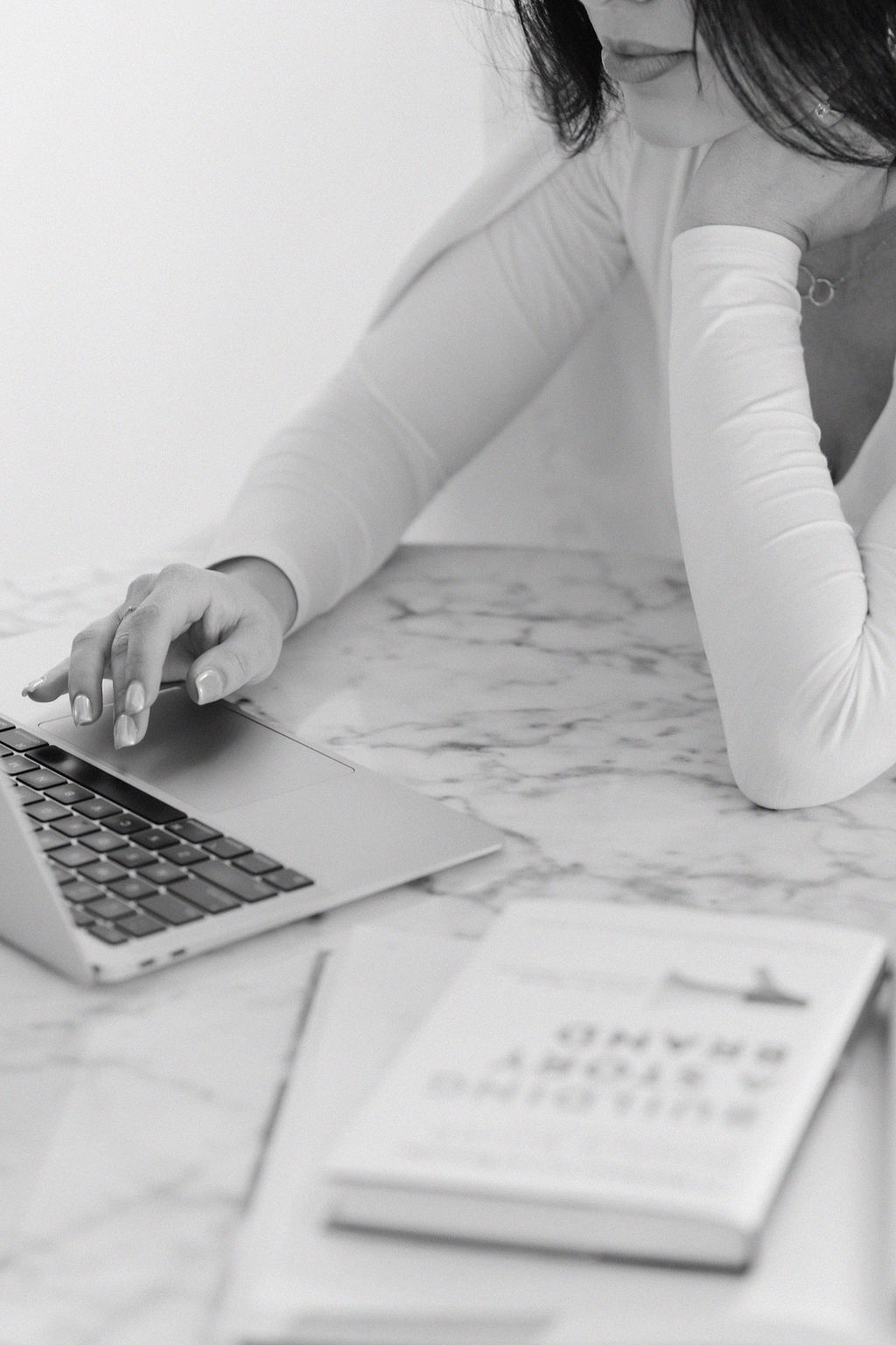 Black and white close up detail of woman working on a laptop with the book 'Building a Storybrand' in front of her on the table