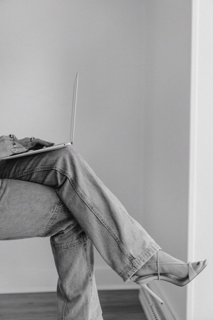 Black and white close up of a pair of crossed legs wearing jeans and strappy stilettos with a laptop perched on top