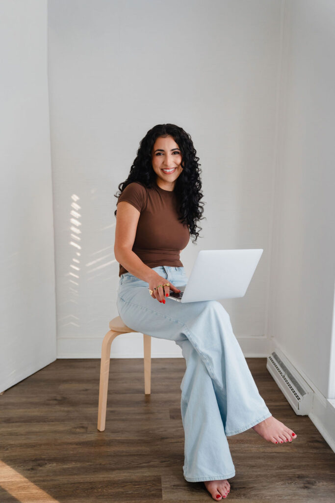 Female email marketing small business owner sits on a wooden stool with a macbook on her lap inside an empty photo studio